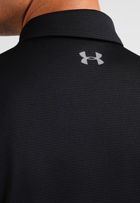 Polo negro con una superficie texturizada, que presenta un logo gris de Under Armour en el cuello de la parte posterior.
