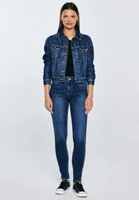 Naine seisab sirgelt, seljas tumesinise teksajope ja teksapüksid, must topp ja mustad tossud, ühevärvilise heleda tausta ees.