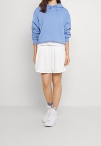 Mladá osoba v světle modré mikině s kapucí, bílé plisované sukni, ponožkách s batikovaným vzorem a bílých kotníkových teniskách stojící na jednobarevném pozadí.