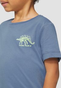 Blauwe katoenen T-shirt met een groene dinosaurus-skelet graphic op de borst. Korte mouwen met een standaard ronde hals.