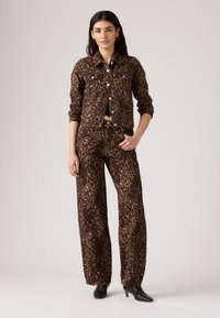 Leopardprint spijkerjack en wijde broek in bruine en zwarte tinten, met knoopsluitingen en voorzakken, gecombineerd met zwarte schoenen.