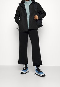 Chaqueta negra impermeable, parte superior de cremallera azul claro, pantalones anchos negros y zapatillas negras con acentos azules. Presenta puños y cremalleras ajustables.