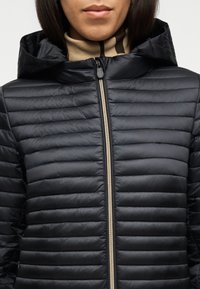 Γυναίκα που φοράει μαύρο καπιτονέ μπουφάν με κουκούλα και εμφανή ασημένιο φερμουάρ, σε συνδυασμό με μπεζ πουλόβερ με ζιβάγκο.