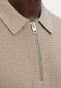 Gros plan d'une personne à la peau foncée portant un haut en maille texturée beige avec un col et une fermeture éclair argentée partiellement fermée.