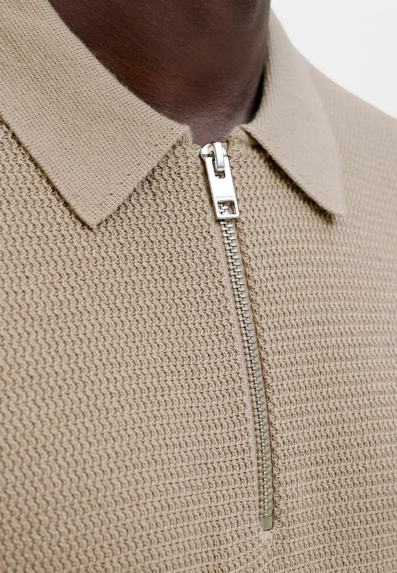 Gros plan d'une personne à la peau foncée portant un haut en maille texturée beige avec un col et une fermeture éclair argentée partiellement fermée.