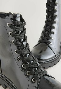 Botines de charol negros con gruesos cordones negros, punta redonda y suela gruesa con relieve. Presentan ojales plateados y paneles texturizados.