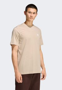 Béžové tričko s krátkym rukávom vyrobené z mäkkej látky, s bielym logom na ľavej strane hrudníka, v kombinácii s tmavohnedými nohavicami.