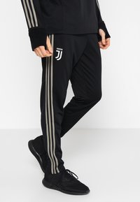 Schwarze Sporthosen mit drei vertikalen dunkelgrauen Streifen und einem Juventus-Logo am linken Bein. Das Material wirkt glatt und sportlich.
