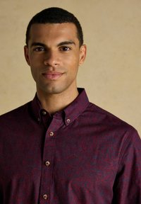 Vīrietis ar īsiem matiem, kas valkā pogātu bordo paisley krekli, skatās uz priekšu uz neitrāla bēša fona.