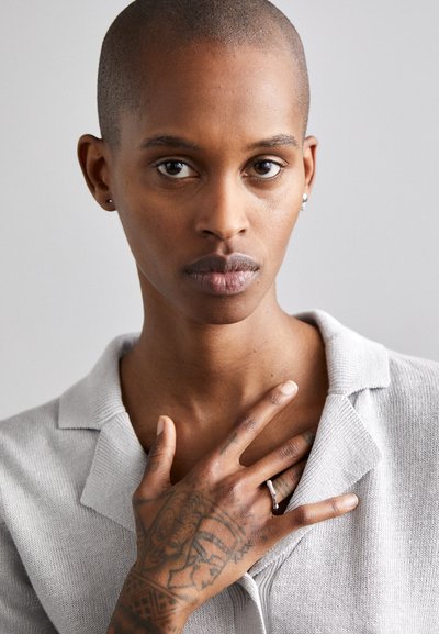 Person with a shaved head wearing a light gray collared sweater, displaying a tattooed hand with a silver ring, against a neutral background.