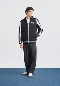 Νέος άντρας που στέκεται σε μπλε πάτωμα, φοράει μαύρο και λευκό μπουφάν Puma με φερμουάρ, μαύρο φαρδύ παντελόνι, λευκά αθλητικά παπούτσια, κοιτάζει προς το πλάι.