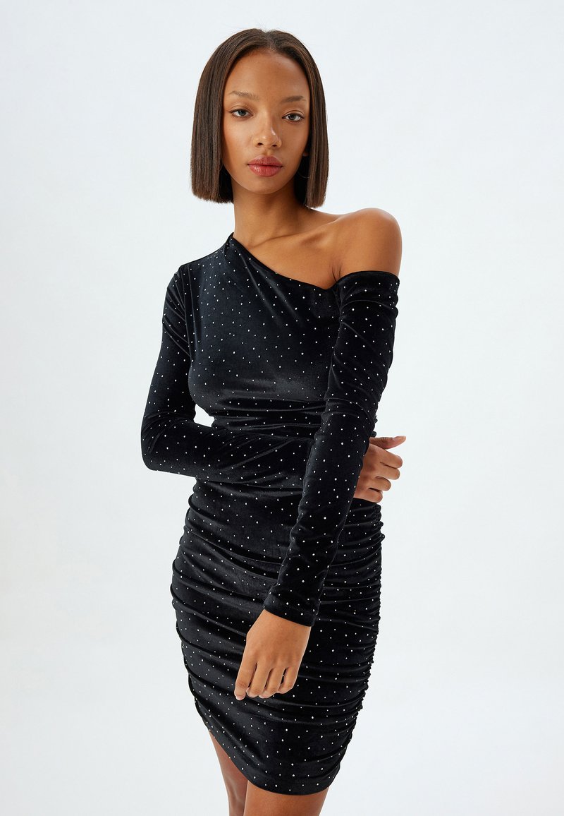 Robe en velours noir avec une seule longue manche, design froncé et embellissements en points blancs éparpillés partout. Texture lisse, forme ajustée.
