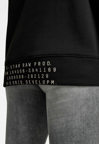 Sudadera negra con ribete acanalado que presenta detalles en texto blanco, combinada con jeans de mezclilla grises. El material parece suave y tiene un diseño casual.