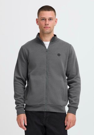 Gray zip-up sweatshirt with a high collar, featuring a small black logo on the chest and side pockets. Made of soft, textured fabric.