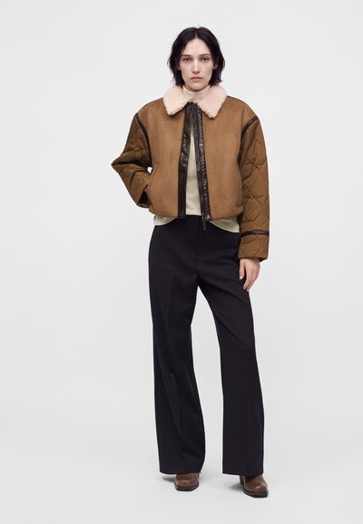 Νεαρή γυναίκα φοράει καφέ shearling μπουφάν με ραμμένα μανίκια, μαύρο παντελόνι με φαρδιά πόδια και καφέ παπούτσια, στέκεται με το ένα χέρι στην τσέπη.