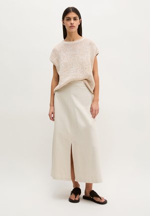 Femme debout portant un haut sans manches beige en tricot, une jupe midi crème avec une fente à l'avant, et des sandales noires sur un fond clair uni.