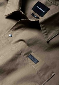 Khaki košeľa na zapínanie na gombíky z hladkej bavlnenej zmesi, ktorá má náprsné vrecko s čiernou nálepkou a kontrastné švy.
