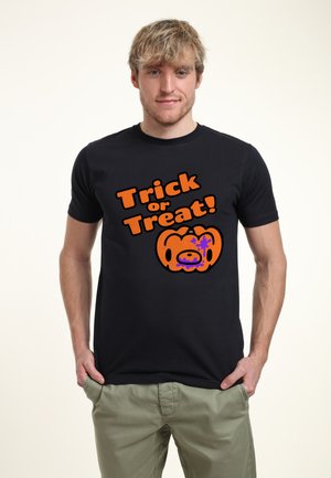 Uomo che indossa una maglietta nera con testo arancione "Trick or Treat!" e una grafica di zucca spaventosa, in piedi con le mani nelle tasche.