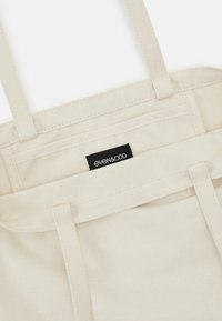 Gros plan sur un sac fourre-tout en toile beige avec deux anses et une étiquette noire de la marque Even&Odd cousue à l'intérieur.