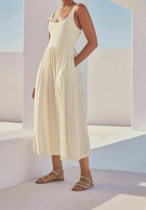 Crèmekleurig jumpsuit met een aansluitend lijfje en wijde pijpen, gecombineerd met lichtbruine sandalen met bandjes en gouden kettingaccenten.