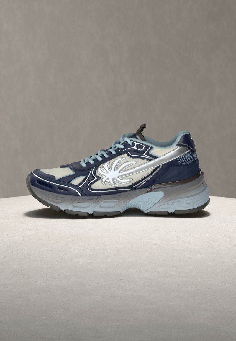 Μπλε και γκρι αθλητικό παπούτσι με λευκό σχέδιο φλόγας στο πλάι, γαλάζια κορδόνια και χοντρή υφή στη σόλα, σε ουδέτερη επιφάνεια και φόντο.
