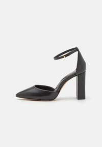 Ballerine en cuir noir avec un bout pointu, une lanière à la cheville et un talon carré, présentant une texture lisse et un design minimaliste.