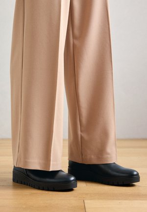 Schwarze Slipper mit glatter Oberfläche und dicker Gummisohle, kombiniert mit beigen, weit geschnittenen Hosen, vor einem Holzfußboden.