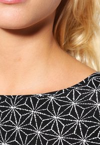 Svart tyg med ett vitt geometriskt mönster, med en bred halsringning. Slät textur, minimal detaljering synlig vid kanten.