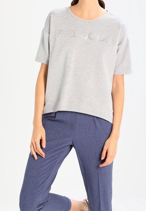 Femme portant un haut gris clair à manches courtes et un pantalon bleu sur mesure, un genou plié, debout devant un fond blanc uni.
