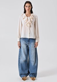 Donna che indossa una blusa crema con colletto arricciato, jeans ampi blu sbiaditi e scarpe dorate a punta con dettagli a fiocco su sfondo semplice.