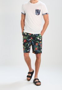 Uomo che indossa una maglietta bianca con taschino e maniche floreali, abbinata a pantaloni corti neri con motivi floreali e sandali neri con fibbia, con le mani in tasca.