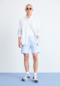 Model iført en hvid og blå stribet skjorte over en hvid tanktop, parret med lyseblå shorts og sportssko. Solbriller tilføjet.