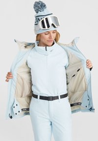 Lichtblauwe skijas met een ritsopening, beige voering en zwarte riem. Draagt over een bijpassende lichtblauwe top. Witte skibril en een blauwe gebreide beanie.