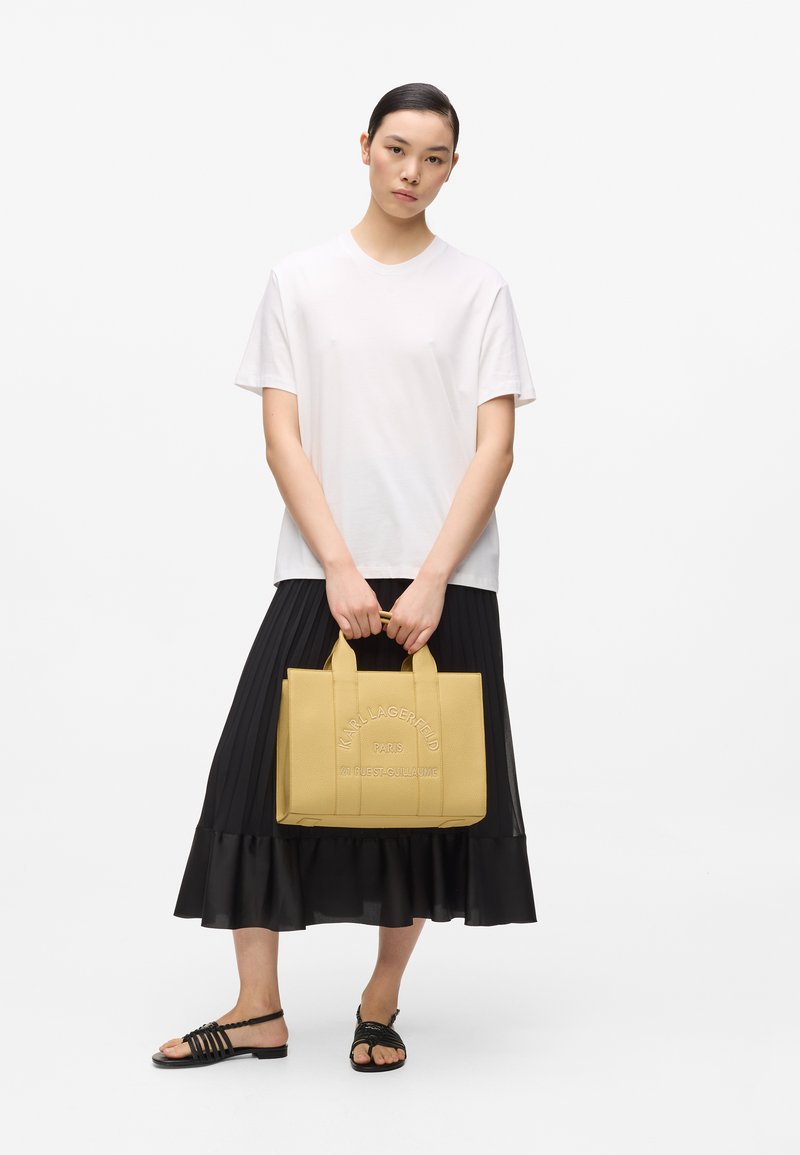 Femme aux cheveux courts et foncés portant un t-shirt blanc et une jupe plissée noire, tenant un sac cabas jaune Karl Lagerfeld et des sandales noires.