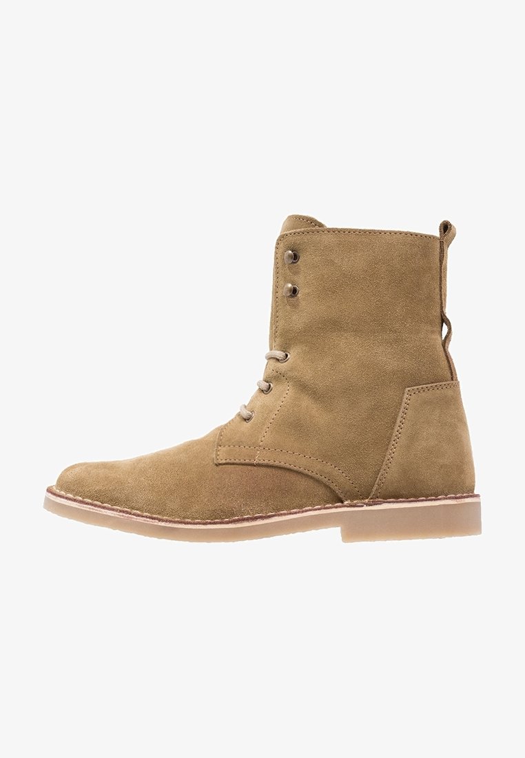 Bottine en daim beige avec lacets à l'avant, œillets métalliques, bout arrondi, semelle plate en caoutchouc et détails cousus sur le côté et la semelle.
