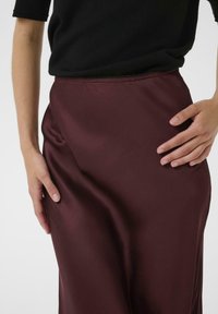 Burgunderfarbene Satin-Rock mit glatter Textur und fließendem Design, ausgestattet mit einem taillierten Bund und dezentem Glanz. Hand auf dem Stoff positioniert.