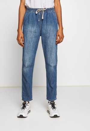 Person iført blå denim joggerbukser med hvid knytesnor og hvide sneakers med sorte snørebånd, stående mod en ensfarvet baggrund.