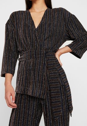 Femme portant un haut cache-cœur noir et un pantalon à rayures verticales multicolores, manches trois-quarts, avec une large ceinture en tissu nouée à la taille.