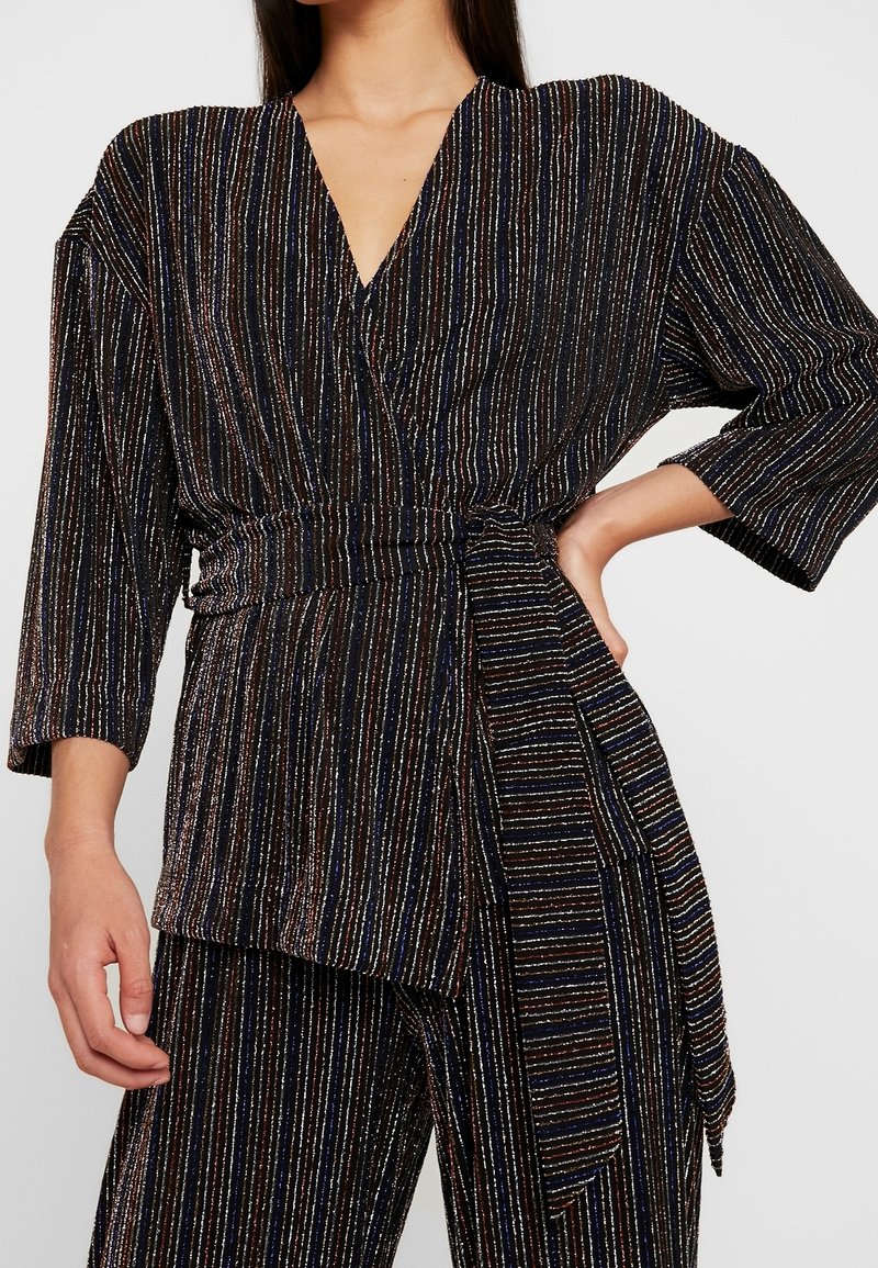 Femme portant un haut cache-cœur noir et un pantalon à rayures verticales multicolores, manches trois-quarts, avec une large ceinture en tissu nouée à la taille.