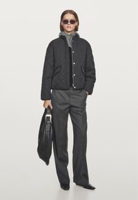 Quilted black bomber jacket with snap buttons, gray collar, and pockets. Paired with gray trousers and holding a black leather bag.