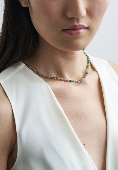 Tuvumā redzama sieviete, kas valkā bezpalsošu baltu augšu un ap viņas kaklu ir krāsains pērļu kaklarota.