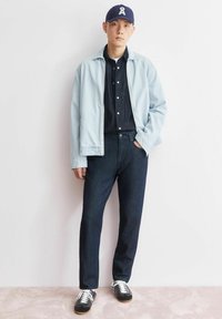 Giacca di jeans azzurro chiaro sopra una camicia di jeans scuro, abbinata a pantaloni scuri a righe e sneakers nere con lacci bianchi. Cappellino con logo.