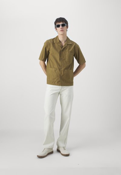 Kurzärmliges olivgrünes Hemd mit zwei vorderen Taschen und Knopfverschluss; kombiniert mit weißen Hosen und cremefarbenen Sneakers. Model trägt eine dunkle Sonnenbrille.