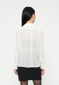 Woman facing away wearing a sheer white long-sleeve blouse with gold button cuffs and a black skirt against a plain white background.