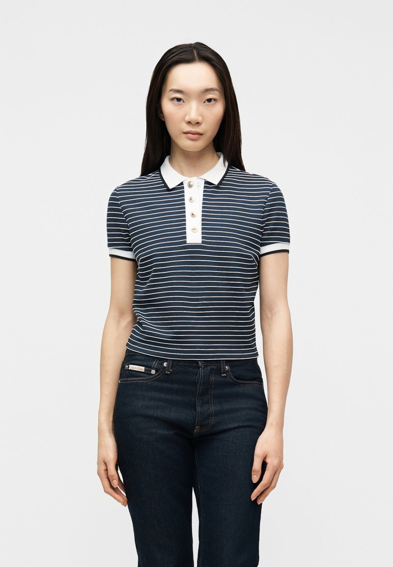 Γυναίκα που φοράει μπλε ναυτικό και άσπρη ριγέ πόλο μπλούζα με λευκά κουμπιά και σκούρο μπλε τζιν, στέκεται μπροστά σε μια απλή ανοιχτόχρωμη επιφάνεια.