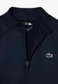 Sudadera azul marino de media cremallera, con tejido texturizado, cuello de canalé y un pequeño logo verde de Lacoste en el lado izquierdo del pecho. Detalle de cremallera destacado.