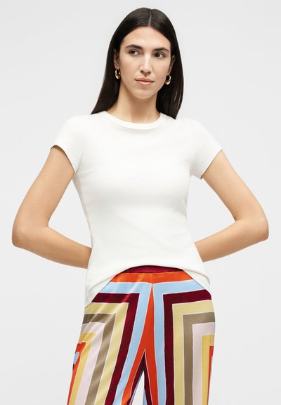 Γυναίκα με μακριά σκούρα μαλλιά που φοράει χρυσούς κρίκους στα αυτιά, λευκό κοντομάνικο τοπ και πολύχρωμο παντελόνι με γεωμετρικό μοτίβο, στέκεται με τα χέρια πίσω από την πλάτη.