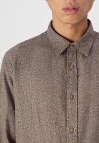 Καφέ, υφασμάτινο πουκάμισο με γιακά και μπεζ κουμπιά, που διαθέτει ένα διακριτικό σχέδιο σε όλη την επιφάνεια του υφάσματος.