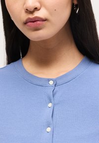 Γυναίκα που φορά ανοιφτό μπλε ριμπ πουκάμισο με κουμπιά, με ίσια σκούρα μαλλιά και ασημένια σκουλαρίκια κρίκους, με κλειστά χείλη και ουδέτερη έκφραση.