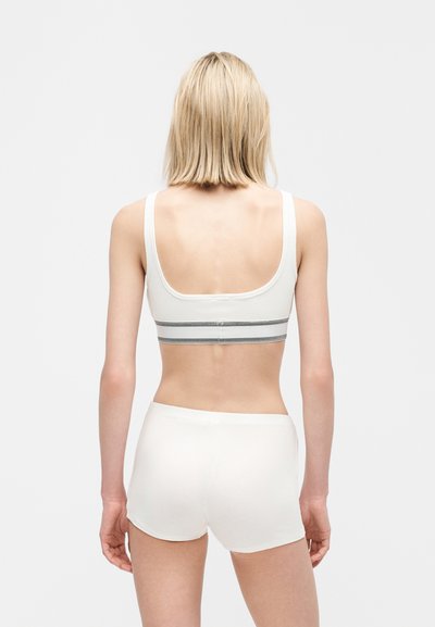 Γυναίκα με κοντά ξανθά μαλλιά, φορώντας λευκό αθλητικό σουτιέν και λευκά σορτς, σε όρθια στάση με την πλάτη προς τα εμπρός, σε απλό φόντο.